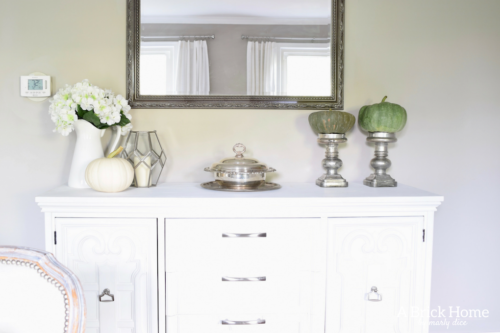 Have YOU seen this gorgeous dining room fall decor with pumpkins and florals? She uses a green and orange color combination to accent her white and silver furniture and decor. Plus, learn how to make this beautiful pumpkin and floral centerpiece. #falldecor #pumpkindecor #falldiningroom