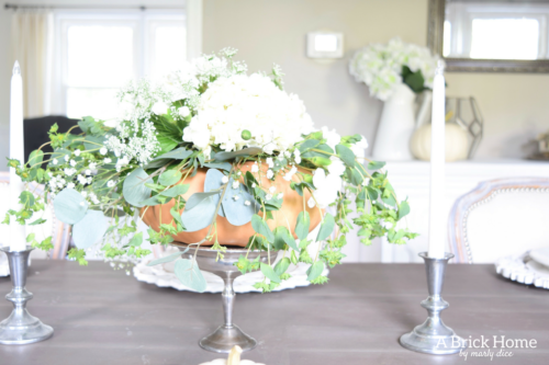 Have YOU seen this gorgeous dining room fall decor with pumpkins and florals? She uses a green and orange color combination to accent her white and silver furniture and decor. Plus, learn how to make this beautiful pumpkin and floral centerpiece. #falldecor #pumpkindecor #falldiningroom