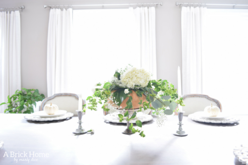 Have YOU seen this gorgeous dining room fall decor with pumpkins and florals? She uses a green and orange color combination to accent her white and silver furniture and decor. Plus, learn how to make this beautiful pumpkin and floral centerpiece. #falldecor #pumpkindecor #falldiningroom